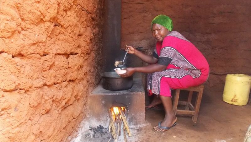 Eine Frau sitzt auf einem hölzernen Hocker und kocht über einem Lehmofen.