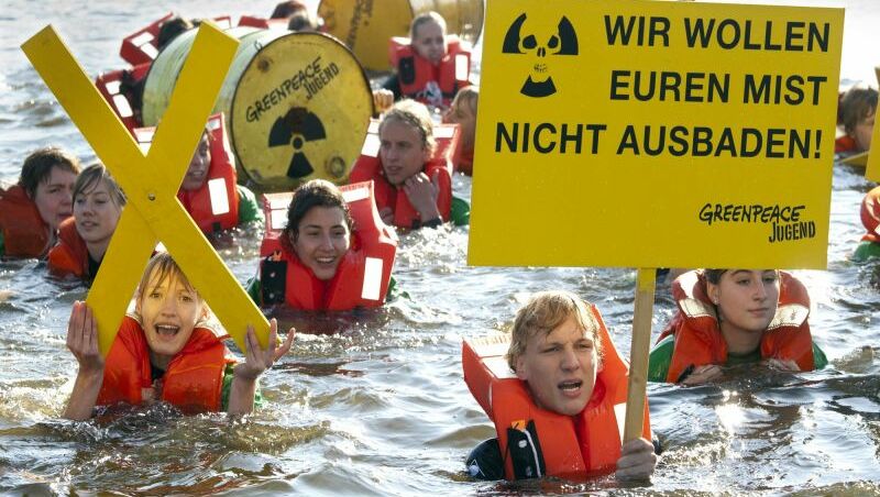 Eine Gruppe Jugendliche steht im Brusttiefen Wasser und hält Protestschilder und bemalte Atommüllkanister hoch. Auf dem Schild im Vordergrund steht: "Wir wollen euren Mist nicht ausbaden! Greenpeace Jugend"