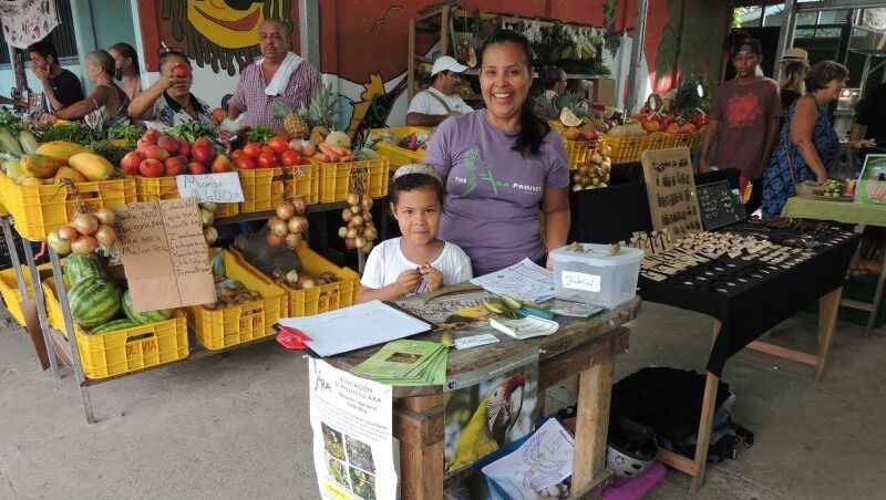 Tirza Morales, Projektleiterin beim Ara Project, steht neben einem jungen Mädchen auf einem Markt mit ihrem Info Stand. Beide lächeln in die Kamera.