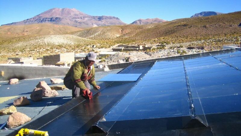 Ein Mann installiert vorsichtig ein Solarpanel. Im Hintergrund ist die hügelige Landschaft der Anden zu sehen.