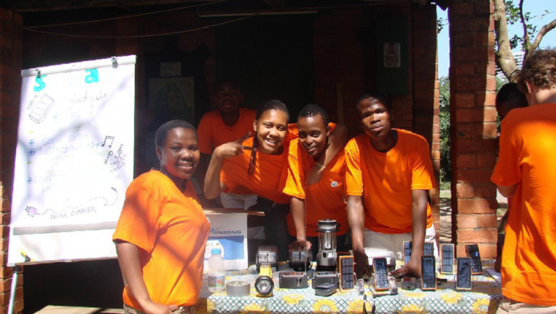 Eine Gruppe Jugendliche steht vor einem Tisch, auf dem Solarenergie Geräte stehen, und lächeln in die Kamera.