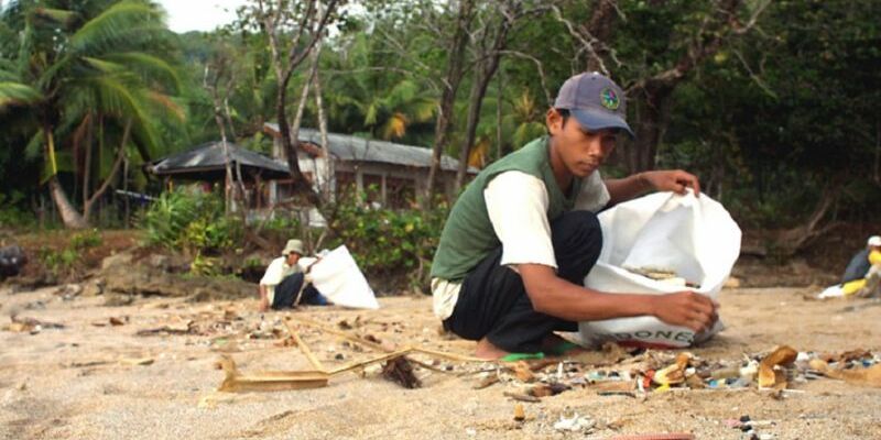 Zwei Jugendliche sammeln Plastikmüll vom Strand auf, im Hintergrund ein Haus und Palmen.