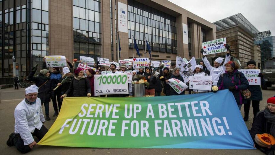 Eine Gruppe Aktivist*innen posieren mit einem großen Banner vor einem EU-Regierungsgebäude. Die Aufschrift des Banenrs: "Serve up a better future for farming!"