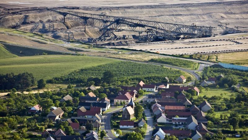 Foto von einem beschaulichen Dorf, umrahmt von grünen Wiesen, im Hintergrund das Kohleabbaugebiet in Jänschwalde.