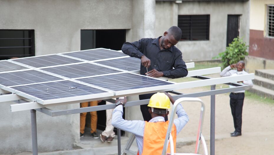 Ein Mann steht auf einer Leiter und schraubt an einem Solarpanel. Im Vordergrund steht ein weiterer Mensch, der eine orangene Warnweste und einen Schutzhelm.