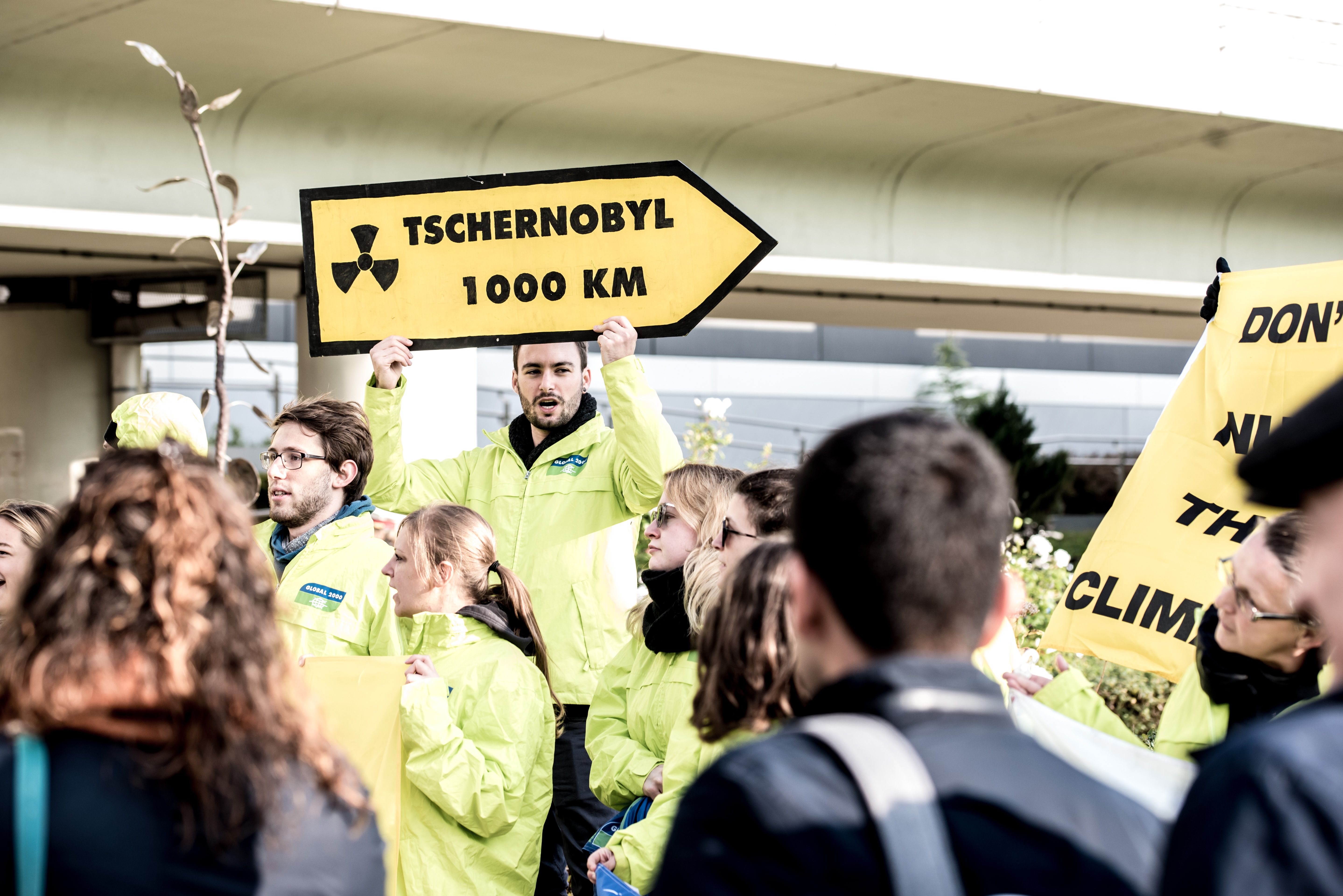 Ein Demonstrant steht in einer Gruppe und hält ein gelb-schwarzes Schild hoch, dass einem Straßenschild ähnelt. Darauf steht: Tschernobyl 1000 km.
