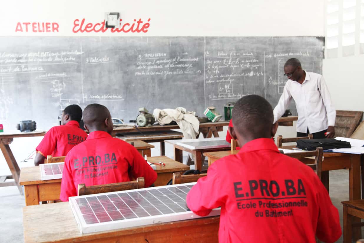 Drei Männer sitzen in einem Klassenzimmer, im Hintergrund eine Kreidetafel. Auf den Tischen vor den Männern liegen Solarpanele. Die Männer tragen alle rote Polohemden mit der Aufschrift "E.PRO.BA"
